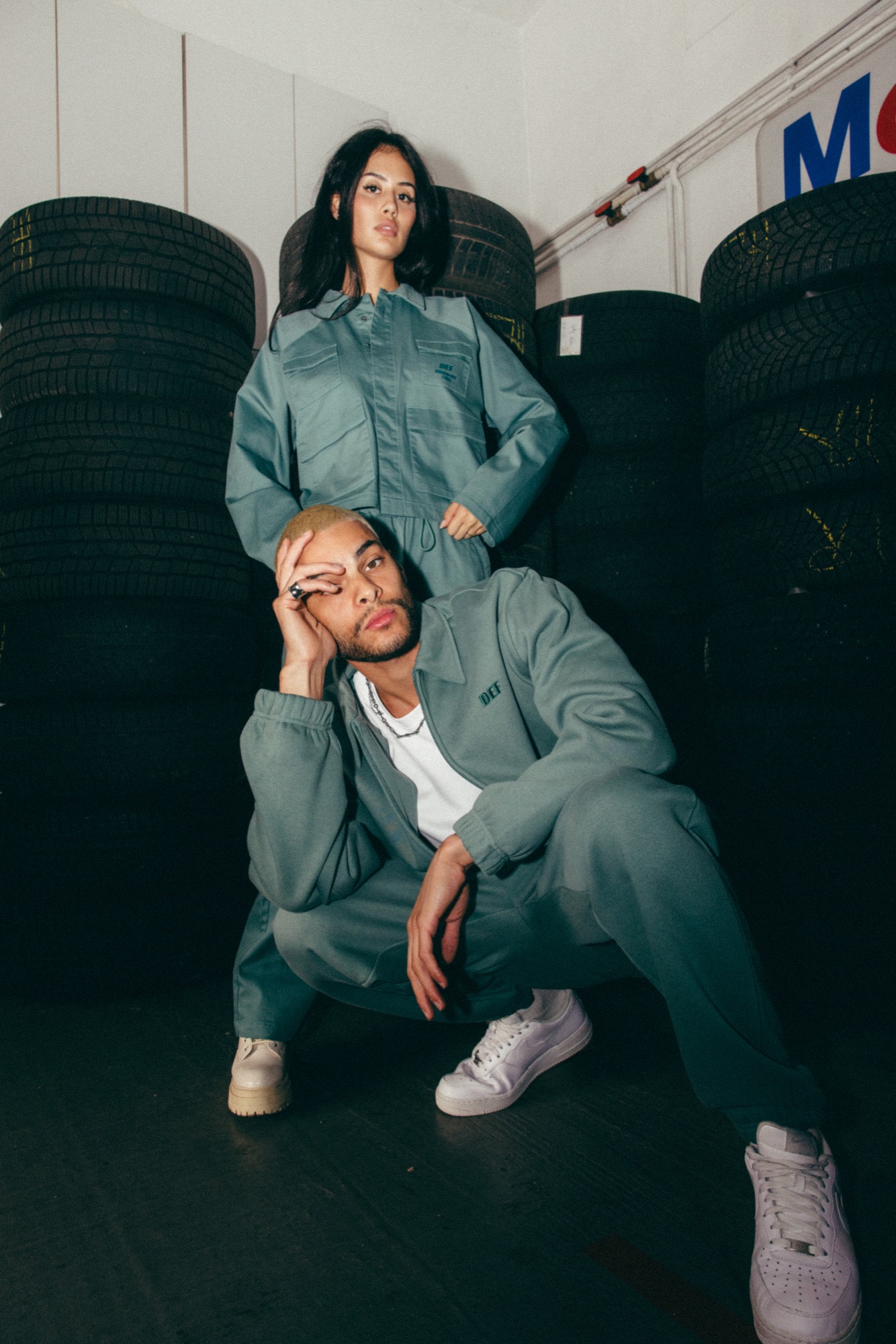 DEFSHOP – Workwear Duo hockend und stehend vor Reifenstapeln