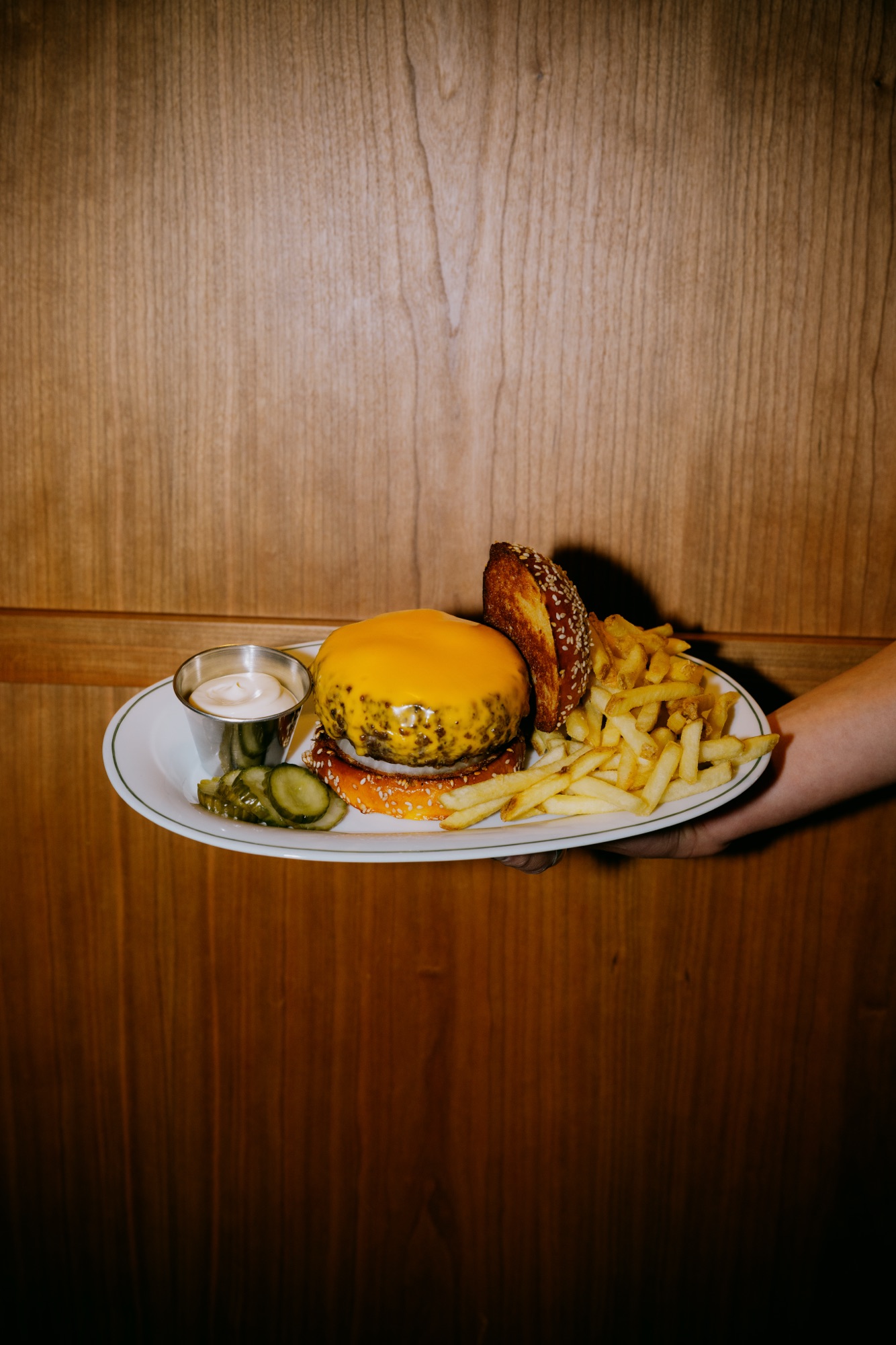 Goldies Bar – Hand hält Teller mit glänzendem Cheeseburger, goldgelben Pommes Frites, Dip und Pickles vor Holzvertäfelung von Kürsat Gürbüz