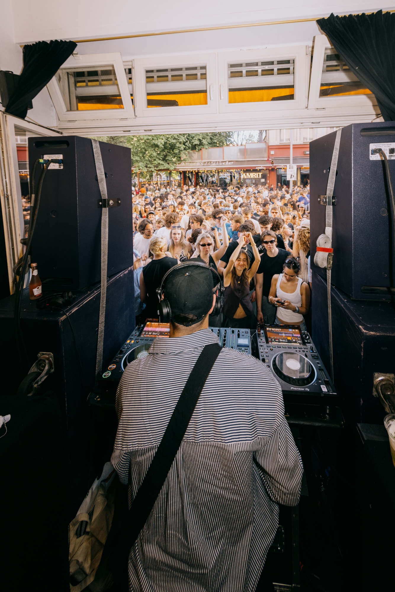 Goldies 1. Mai Fest – Perspektive hinter dem DJ am Mischpult durch das offene Fenster auf die tanzende Straßenmenge von Kürsat Gürbüz