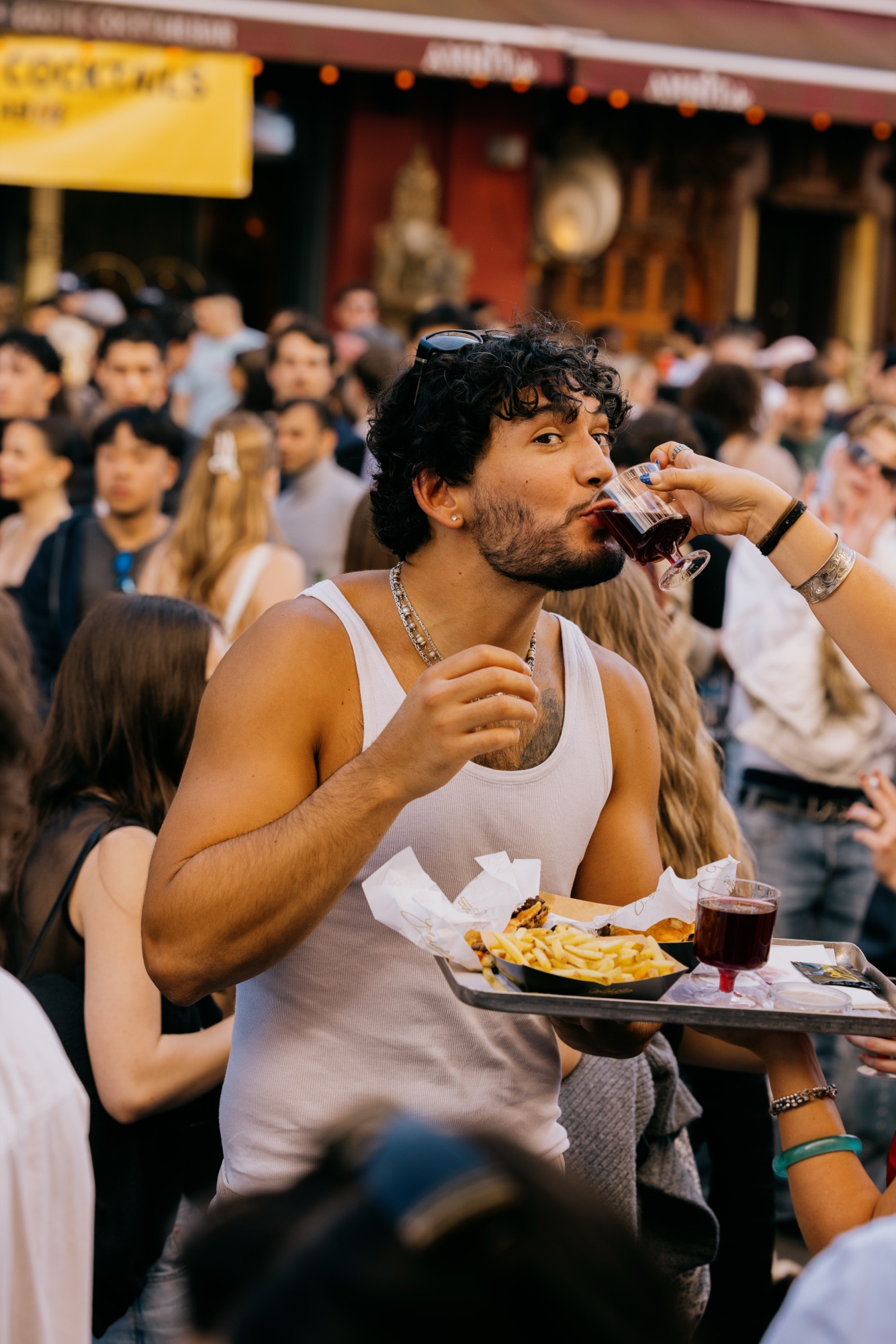 Goldies 1. Mai Fest – Gast trinkt Wein aus einem Glas während er ein volles Tablett mit Burger und Pommes trägt von Kürsat Gürbüz