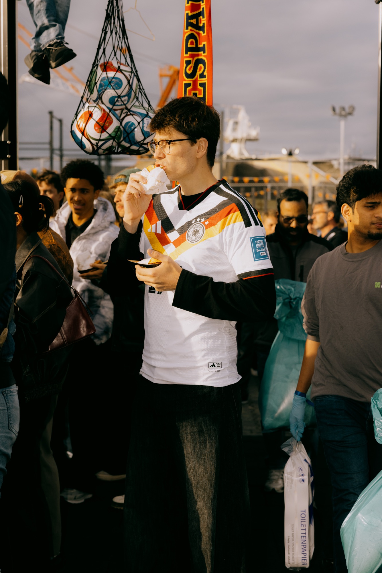 Goldies × Adidas Sportsbase – Fan im Deutschland-Trikot isst Burger unter Ballnetz von Kürsat Gürbüz