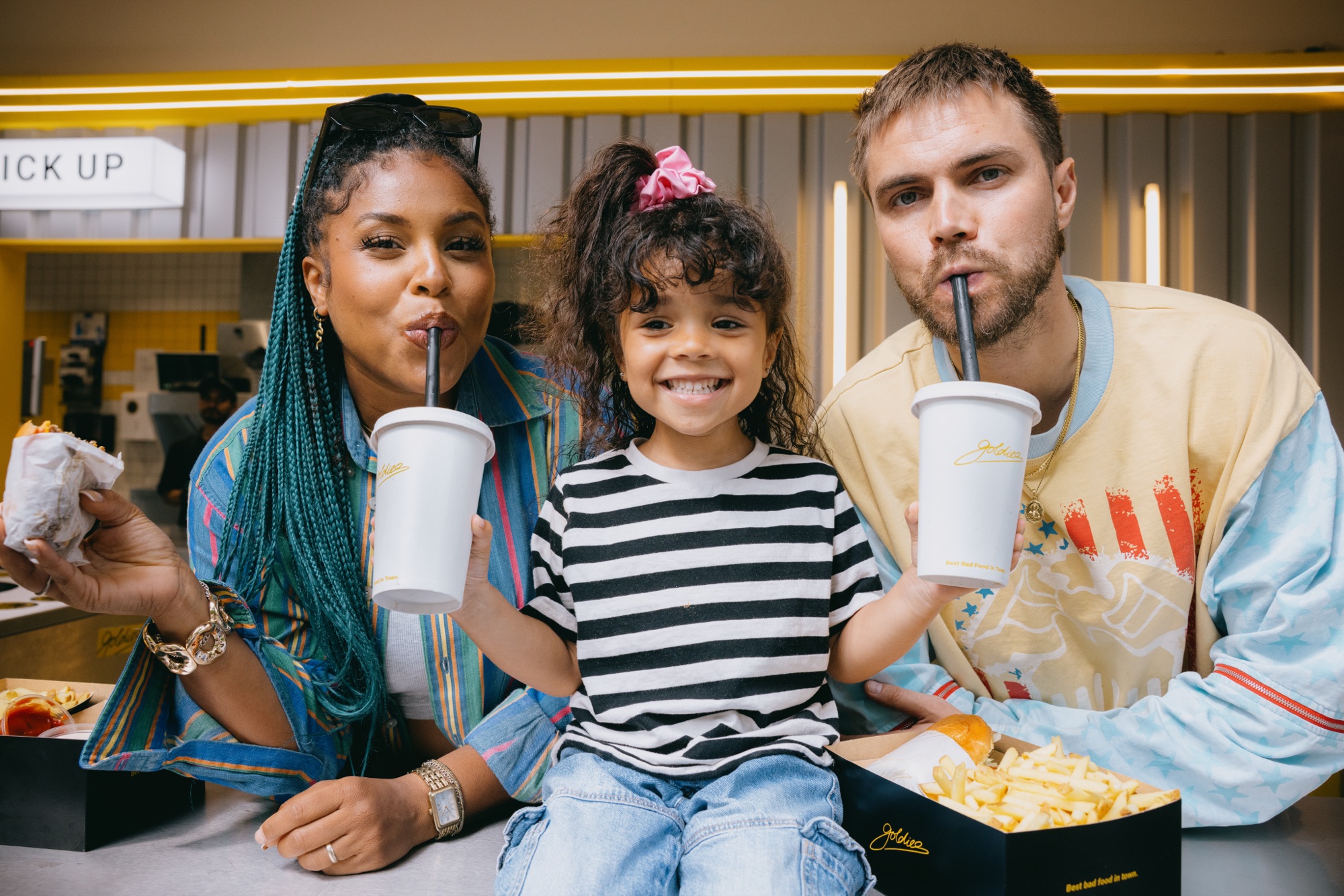 Goldies Kampagne – Familie mit Kind an der Goldies Pick-Up Theke mit Milkshake-Bechern und Pommes-Box von Kürsat Gürbüz