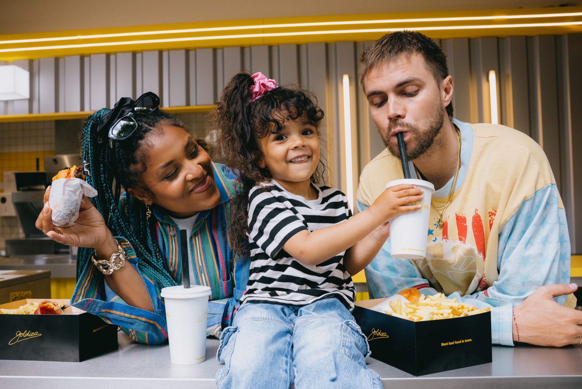 Goldies Kampagne – Familie mit Kind am gelben Tisch beim Teilen von Burger, Pommes und Milkshakes von Kürsat Gürbüz