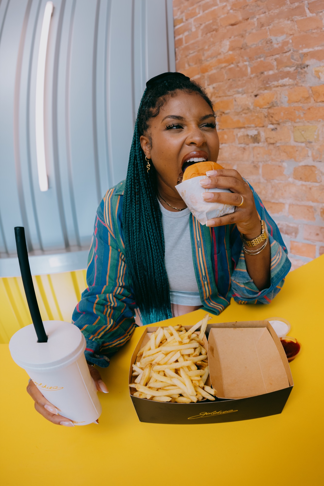 Goldies Kampagne – Porträt einer Frau beim Hineinbeißen in einen Burger am gelben Tisch mit Pommes und Milkshake von Kürsat Gürbüz