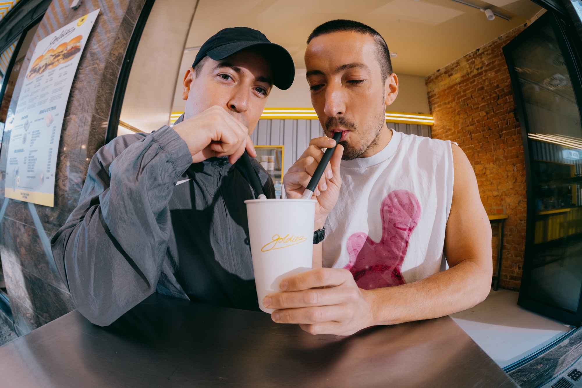 Goldies Kampagne – Fisheye-Szene zweier Freunde, die gemeinsam mit Strohhalmen aus einem Goldies-Milkshake-Becher trinken von Kürsat Gürbüz