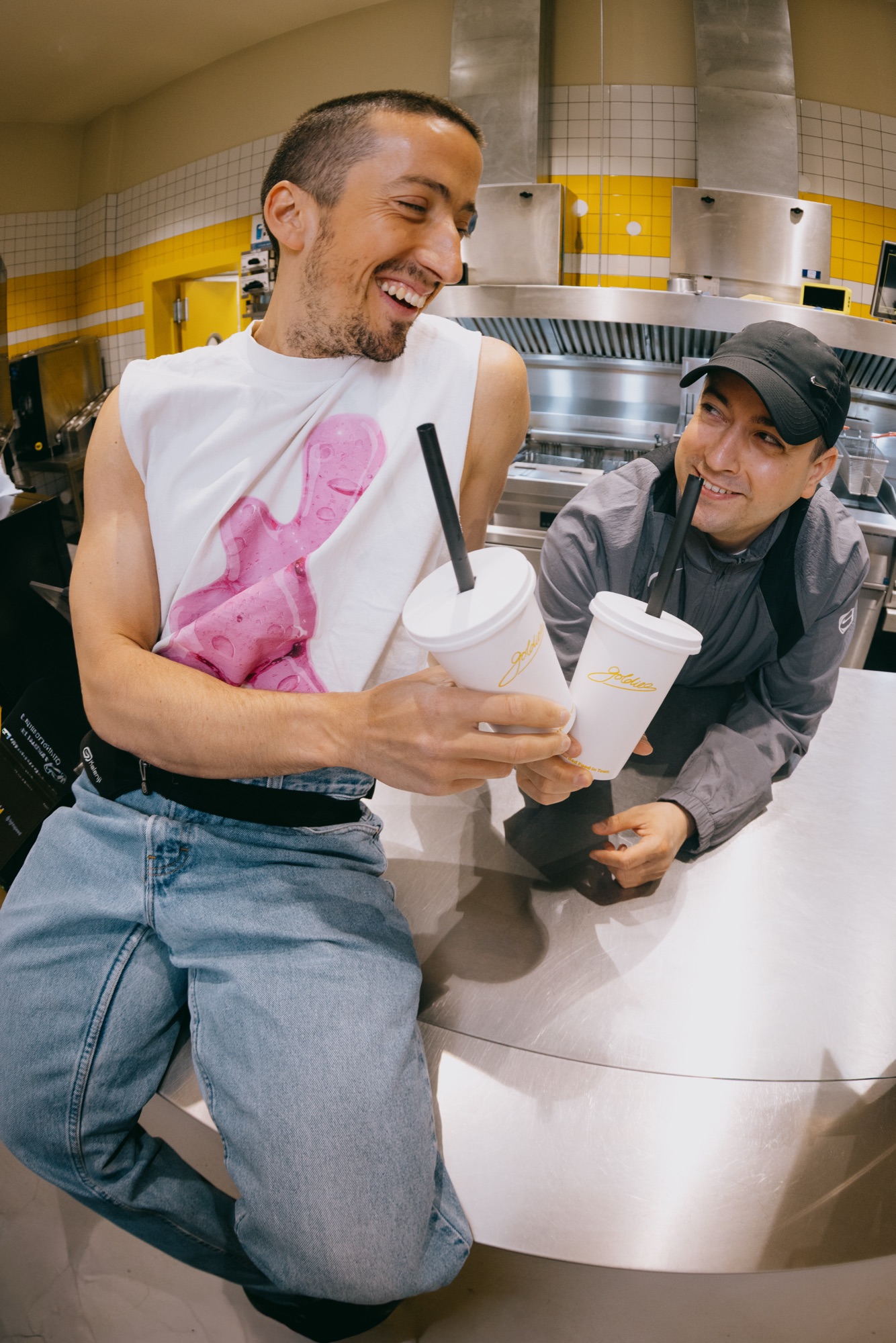 Goldies Kampagne – Zwei Männer lachen an der Edelstahl-Theke der offenen Küche mit Goldies-Milkshake-Bechern von Kürsat Gürbüz