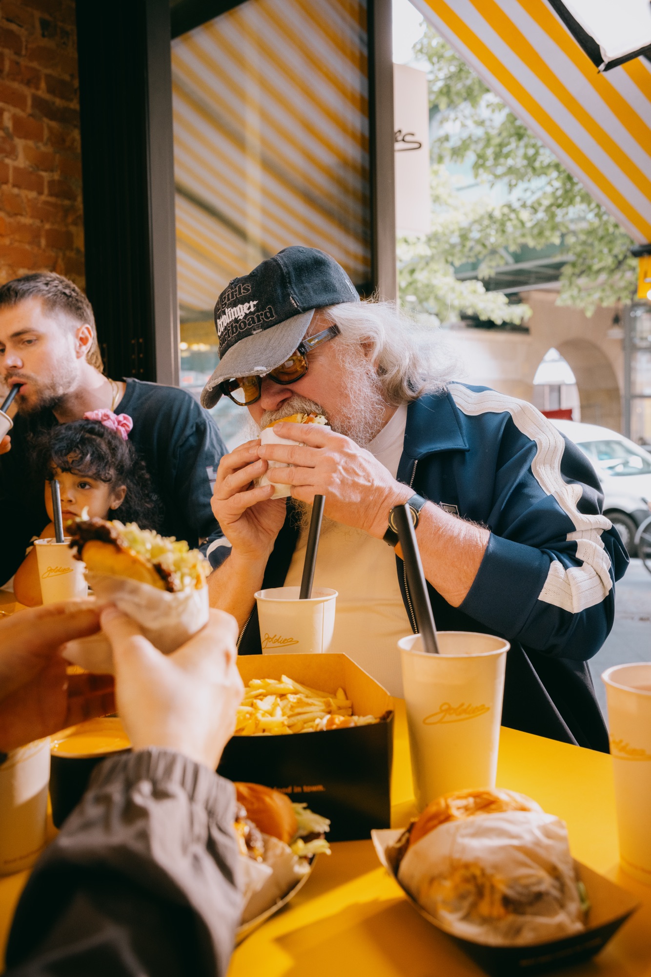 Goldies Kampagne – Älterer Mann mit Sonnenbrille und Cap beißt am gelben Außentisch in einen Smashburger, umgeben von Pommes und Milkshakes von Kürsat Gürbüz
