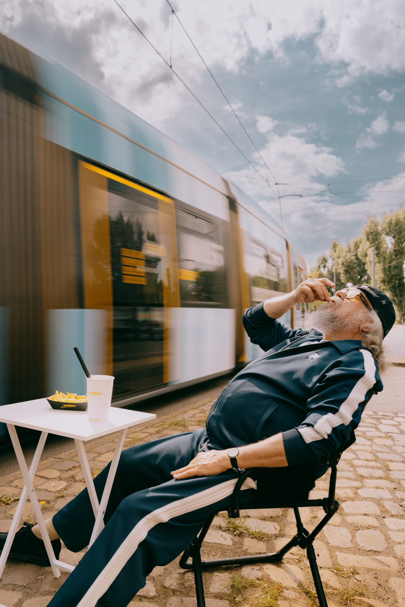 Goldies Kampagne – Älterer Mann entspannt im Klappstuhl am Campingtisch mit Pommes und Milkshake, während eine Berliner Tram mit Bewegungsunschärfe vorbeifährt von Kürsat Gürbüz