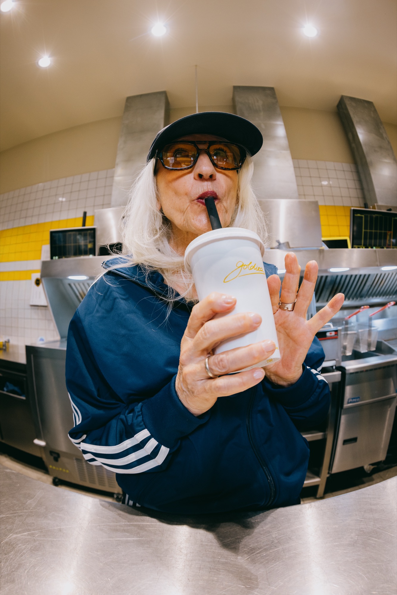 Goldies Kampagne – Fisheye-Porträt einer älteren Frau mit weißem Haar, Sonnenbrille und blauem Trainingsanzug beim Trinken eines Milkshakes in der Küche von Kürsat Gürbüz