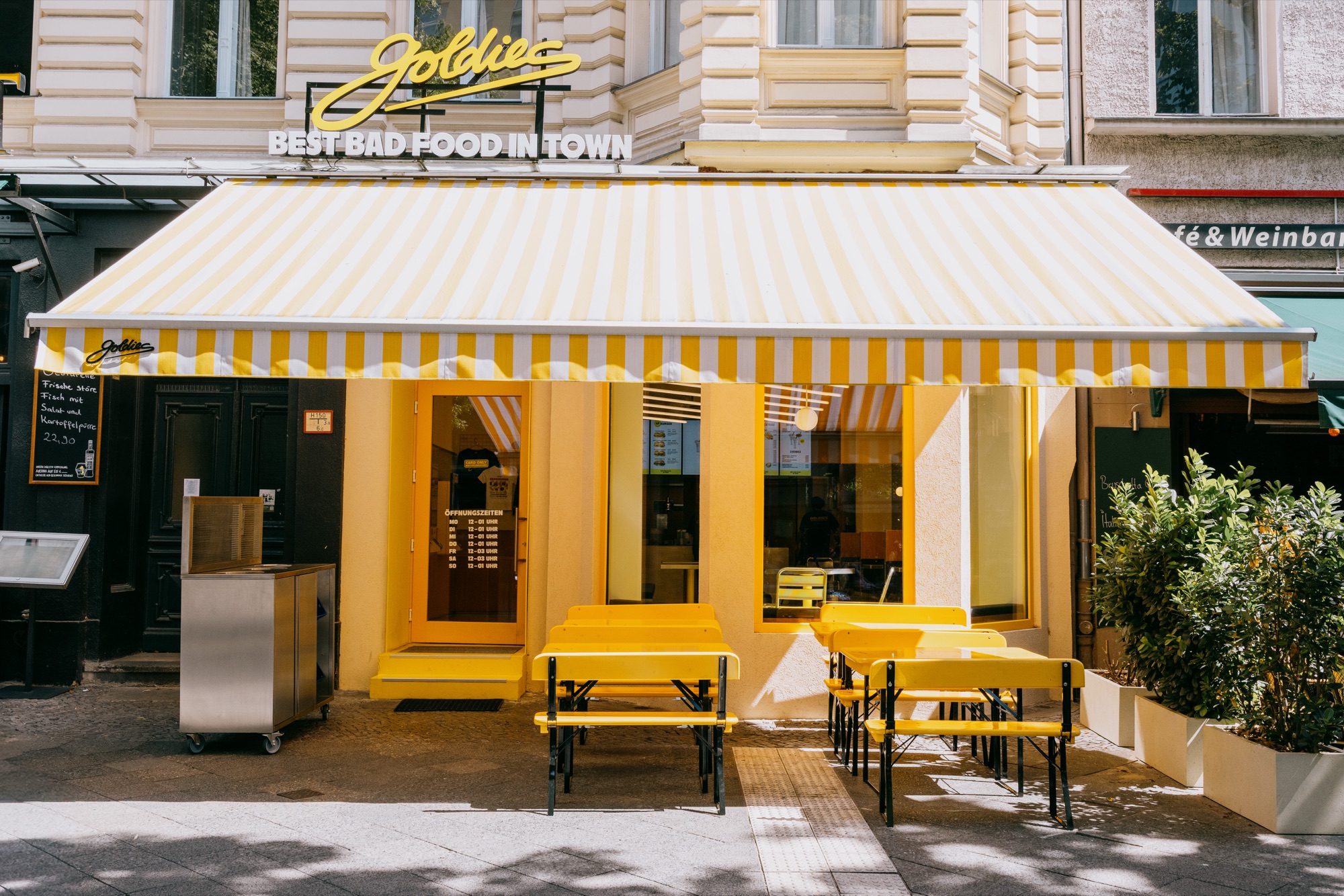 Goldies Schöneberg – Außenfassade mit gelb-weiß gestreifter Markise, Leuchtschrift Best Bad Food in Town und gelben Picknicktischen von Kürsat Gürbüz