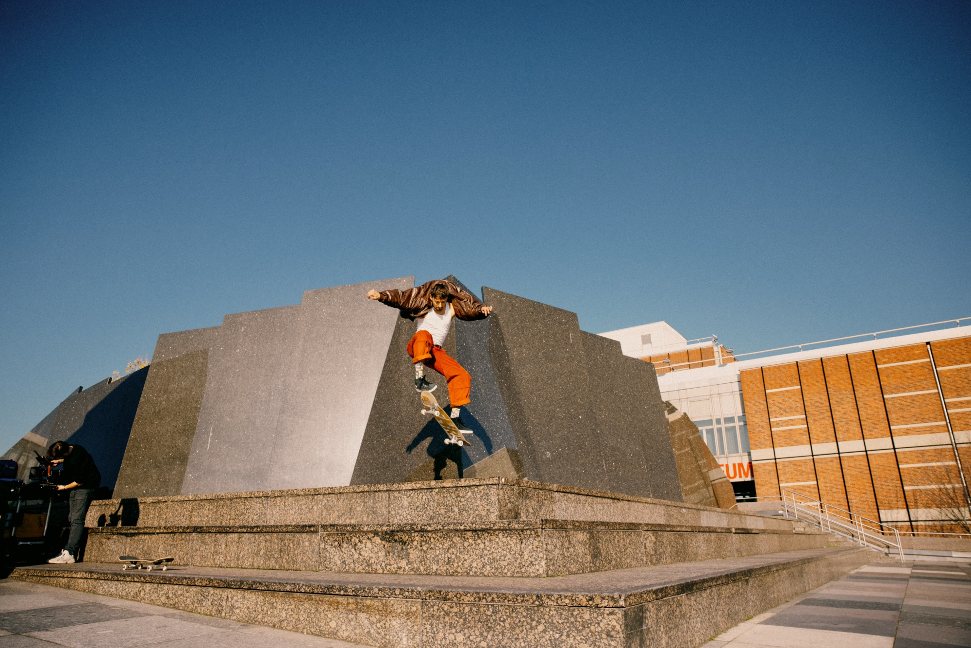 Wiggle Steps – Skater mid-air Sprung über brutalistische Steinstufen gegen blauen Himmel von Kürsat Gürbüz